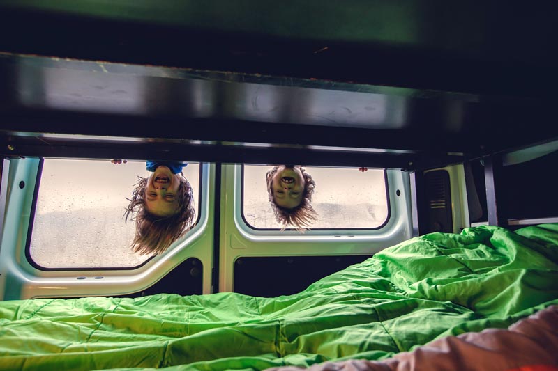 Road trip en famille en van © Tomsickova/stock.adobe.com 2 enfants se tenant sur le lit mezzanine avec leurs têtes à l'envers dans un van aménagé
