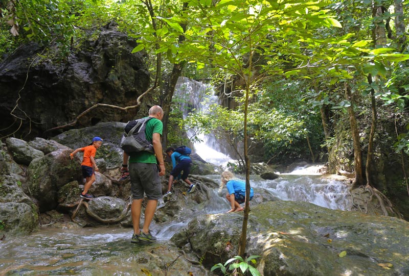 3 enfants et 1 adulte sautant sur les rochers devant une cascade dans la jungle en Tha&iuml;lande