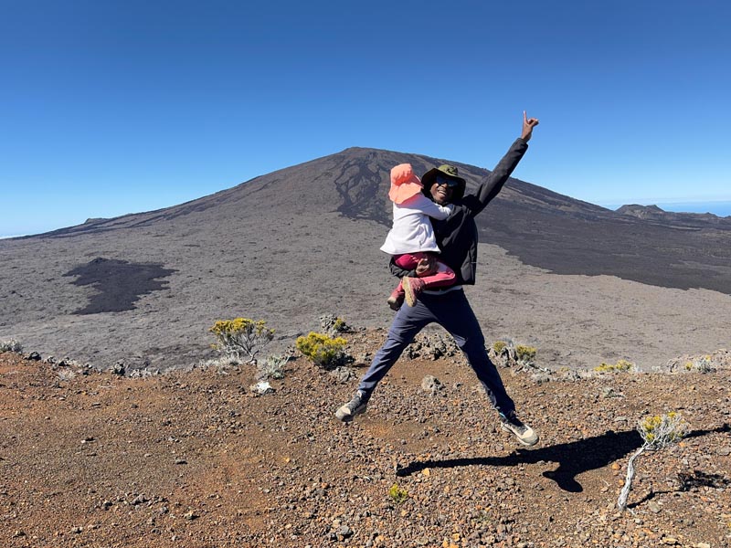 Randonnée au piton de la Fournaise sur l'île de la Réunion © Mylène Mieuze/Nomade Aventure Un père tenant sa fille dans les bras sautant avec le piton de la Fournaise en toile de fond